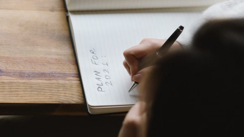 From above of crop unrecognizable female with dark hair sitting at wooden table and writing plan for 2021 in notebook